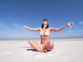 Carefree woman wearing bikini sitting on beach, releasing sand from hand and enjoying