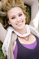Portrait of beautiful blonde woman wearing scarf over top lying in grass, smiling widely