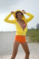 Portrait of pretty woman in bikini, orange shorts and yellow jacket standing on beach