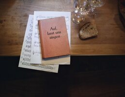 Songbook and music sheet with piece of bread and glass on wooden table