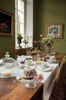 A long breakfast table with cake on a cake stand and under a glass cloche in a a traditional dining room with green-painted walls