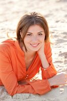 Portrait of pretty brunette woman wearing orange blouse lying on sand, smiling