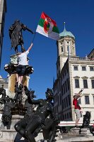 Man holding flag standing on Augustus fountain in front of Perlachturm, Augsburg, Germany