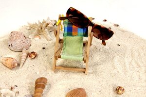 Liegestuhl und Sonnenbrille umgeben von Muscheln im Sand