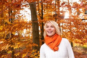 Blonde woman wearing orange scarf and bright sweater smiling in autumn forest