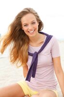 Beautiful blonde woman with windswept hair wearing pink top sitting on beach, smiling