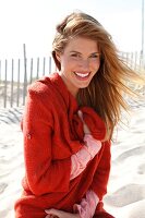 Portrait of attractive woman wearing pink top and red sweater sitting on sand, smiling