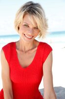 Portrait of pretty blonde woman with short hair wearing red top sitting on beach, smiling