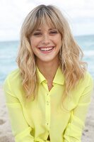 Portrait of cheerful blonde woman wearing yellow shirt sitting on beach, smiling