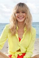 Portrait of cheerful blonde woman wearing yellow shirt sitting on beach, smiling