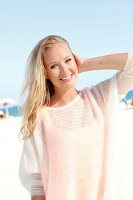 Blonde Frau im weißen Pulli am Strand, lächelt in die Kamera