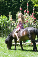 Mädchen in gestreiftem Kleid reitet Pony im sommerlichen Garten