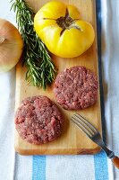Raw Buffalo Burger Patties on a Cutting Board; Tomato, Onion and Rosemary
