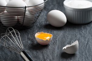 A cracked egg, a whisk and a wire basket of eggs on a stone surface