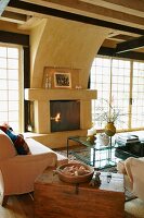 Alte Holztruhe zwischen Couch und Sessel um Glastisch und offener Kamin in Wohnzimmer mit Sprossen Terrassentüren