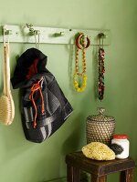 Green bathroom wall with wooden wall coat rack with jewelry hanging from it and a homemade fabric tote bag; underneath an antique side table