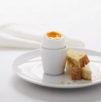 A boiled egg in a white egg-cup with three fingers of lightly toasted bread