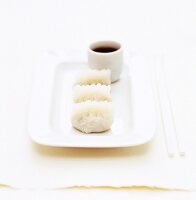 A white plate of three dumplings with a white ramekin of soy sauce dip