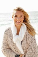 A young blonde woman by the sea wearing a beige knitted cardigan and a scarf