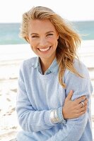 A young blonde woman by the sea wearing a blue knitted jumper and a denim shirt