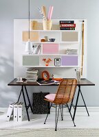 A self-designed home office – a black table in front of a white, mass-produced panel set with shelves and coloured boxes and a retro wicker chair with a black metal frame