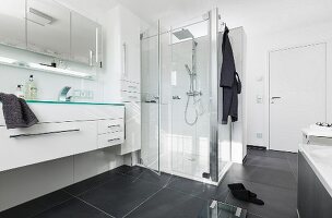 Grosszügiger Waschtisch mit weißem Schrankunterbau, darüber mehrtüriger Spiegelschrank und beleuchtete Glasplatte, im Hintergrund Glas Duschkabine in modernem, schwarzweissem Bad mit schwarzen, grossformatigen Bodenfliesen