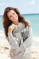 A young brunette woman by the sea wearing a grey sweatshirt and a scarf