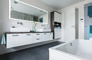 A double washstand with console basins on a drawer unit with a wall-mounted LED mirror above it and the corner of the bathtub in the foreground