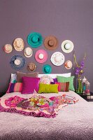 Doppelbett mit verschiedenfarbigen Kissen vor lila Wand und aufgehängter Hutsammlung