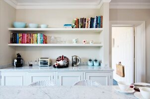 View across marble table top to fitted sideboard in niche below books and ornaments on two white, floating shelves in vintage ambiance