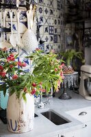 Vintage jug of flowers on kitchen worksurface in front of kitchen utensils hung on wall tiled with blue and white patterned, old-fashioned tiles