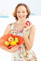 Junge Frau im Sommerkleid hält mit Obst gefüllten Hut und einen Apfel
