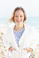 A young blonde woman on the beach wearing a shirt and a short, white jacket