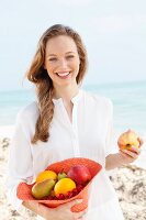 A young woman by the sea wearing a white blouse holding a hat filled with fruit