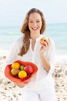 A young woman by the sea wearing a white blouse holding a hat filled with fruit