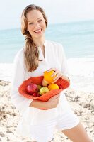 A young woman by the sea wearing a white blouse and shorts holding a hat filled with fruit