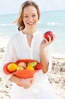 A young woman by the sea wearing a white blouse holding a hat filled with fruit