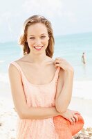 Fröhliche, junge Frau in apricotfarbenem Spitzenkleid und mit Hut in der Hand am Meer
