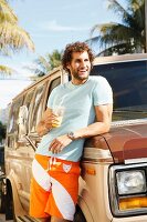A young man with curly hair and a beard wearing summer clothes leaning against a car