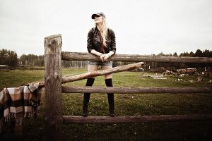 A long-haired, blonde woman standing on a wooden fence wearing a leather jacket and short shorts