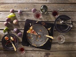 The remains of a smoked ham and feta cheese quiche on a rustic table with fresh figs and glasses of red wine