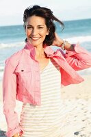 A brunette woman on a beach wearing a short pink denim jacket