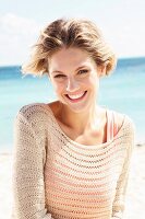 A young blonde woman on a beach wearing a salmon-coloured top and a beige openwork jumper