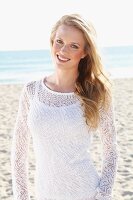 A young blonde woman on a beach wearing a white top and a transparent knitted jumper