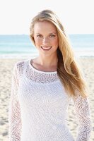 A young blonde woman on a beach wearing a white top and a transparent knitted jumper