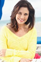 A brunette woman on a beach with her arms folded wearing a yellow knitted jumper
