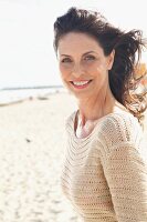 A brunette woman on a beach with her her blowing in the wind wearing a beige, openwork jumper