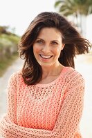 A brunette woman on a beach wearing a salmon-coloured top and an openwork jumper