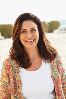 A brunette woman on a beach wearing a white top and colourful cardigan