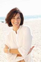 A brunette woman on a beach with her arms folded wearing a thin white blouse